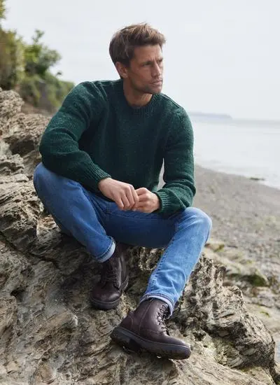 Man sitting on a rock on a beach in a bottle green fisherman flecked crew neck sweater