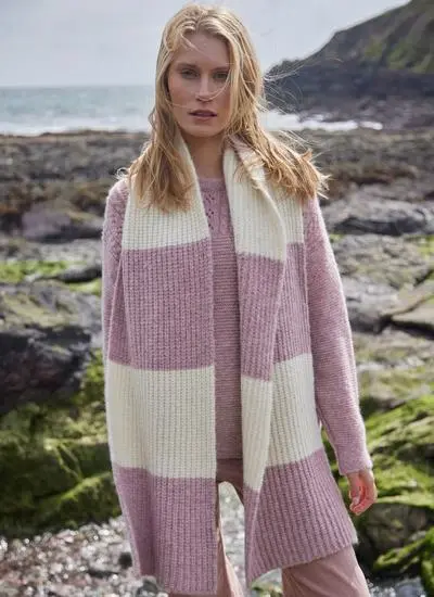 blonde woman standing on rocky, mossy beach wearing large knit pink and white scarf