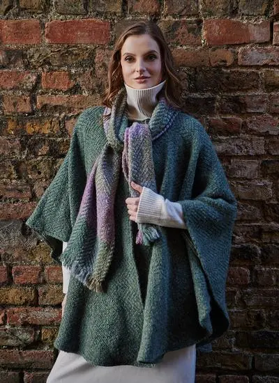Blonde woman standing against red brick wall wearing sage cape with purple scarf.
