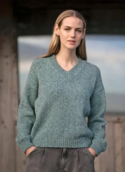 woman with blonde hair standing outside stable with hands in pockets wearing jade fisherman knitted sweater