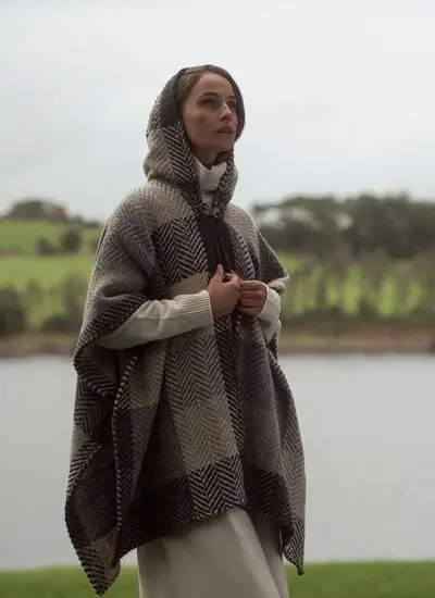 Side angle shot of woman standing in front of lake wearing silk slip dress and beige check cape with hood up.