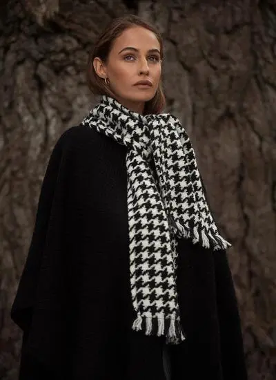 Woman in woodland wearing black wool cape with houndstooth scarf looking at camera.