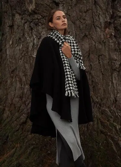 Woman in woodland wearing black wool cape with houndstooth scarf looking up.