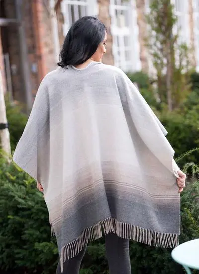 Back angle shot of brunette woman wearing black and beige fringed shawl with building and greenery in background. 
