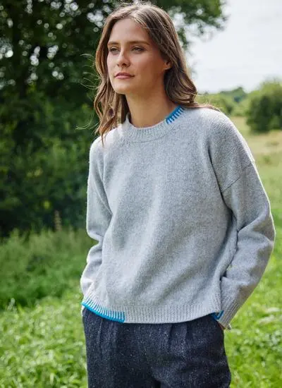 brunette woman standing in park with hands in pockets wearing grey sweater