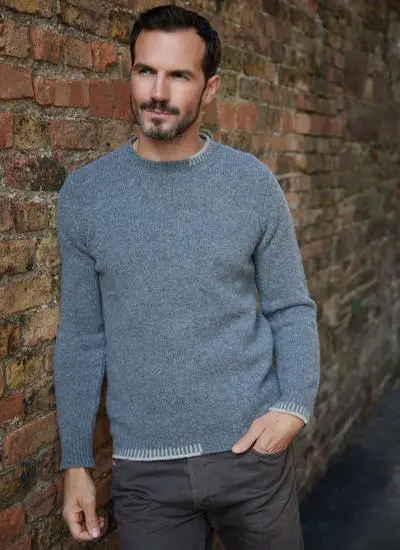 brunette man wearing grey sweater leaning against red brick wall with one hand in pocket