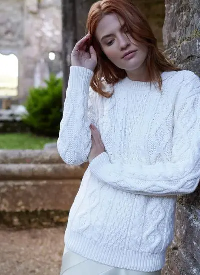 red haired woman leaning against wall in stony passageway with arms folded wearing a white aran sweater