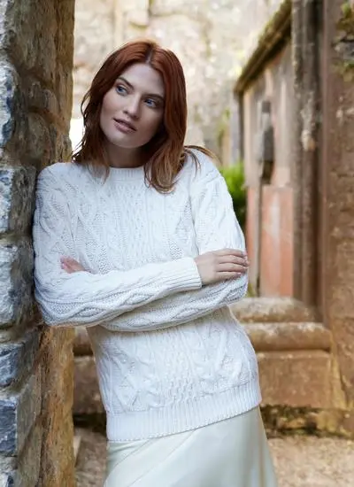 red haired woman leaning against wall in stony passageway with arms folded wearing a white aran sweater