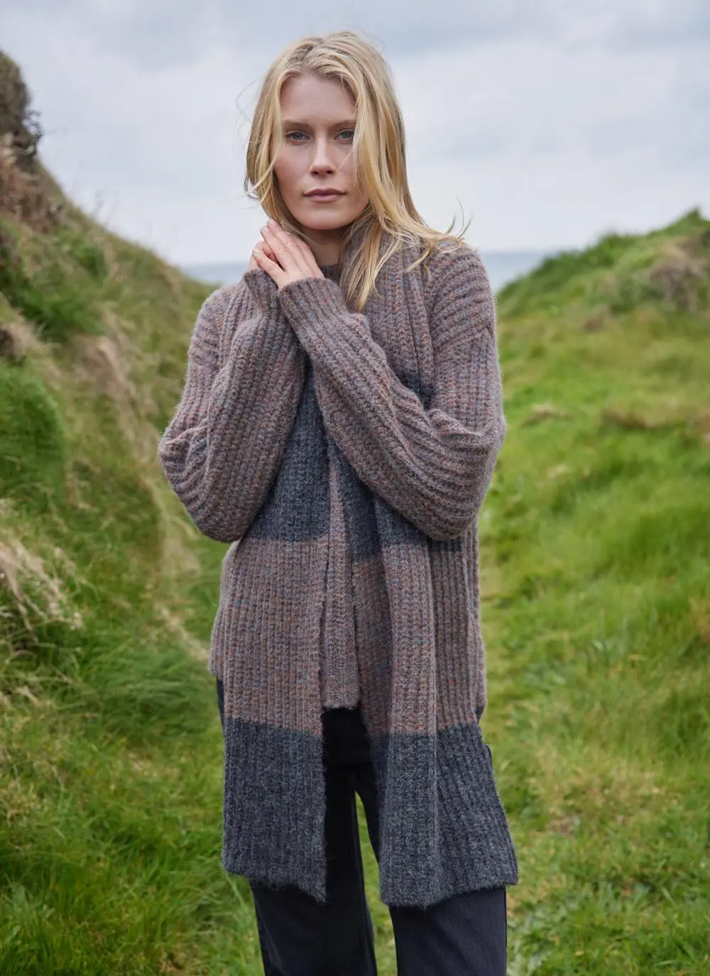 blonde woman standing in grassy coastal area wearing a copper and grey knit scarf with clasped hands