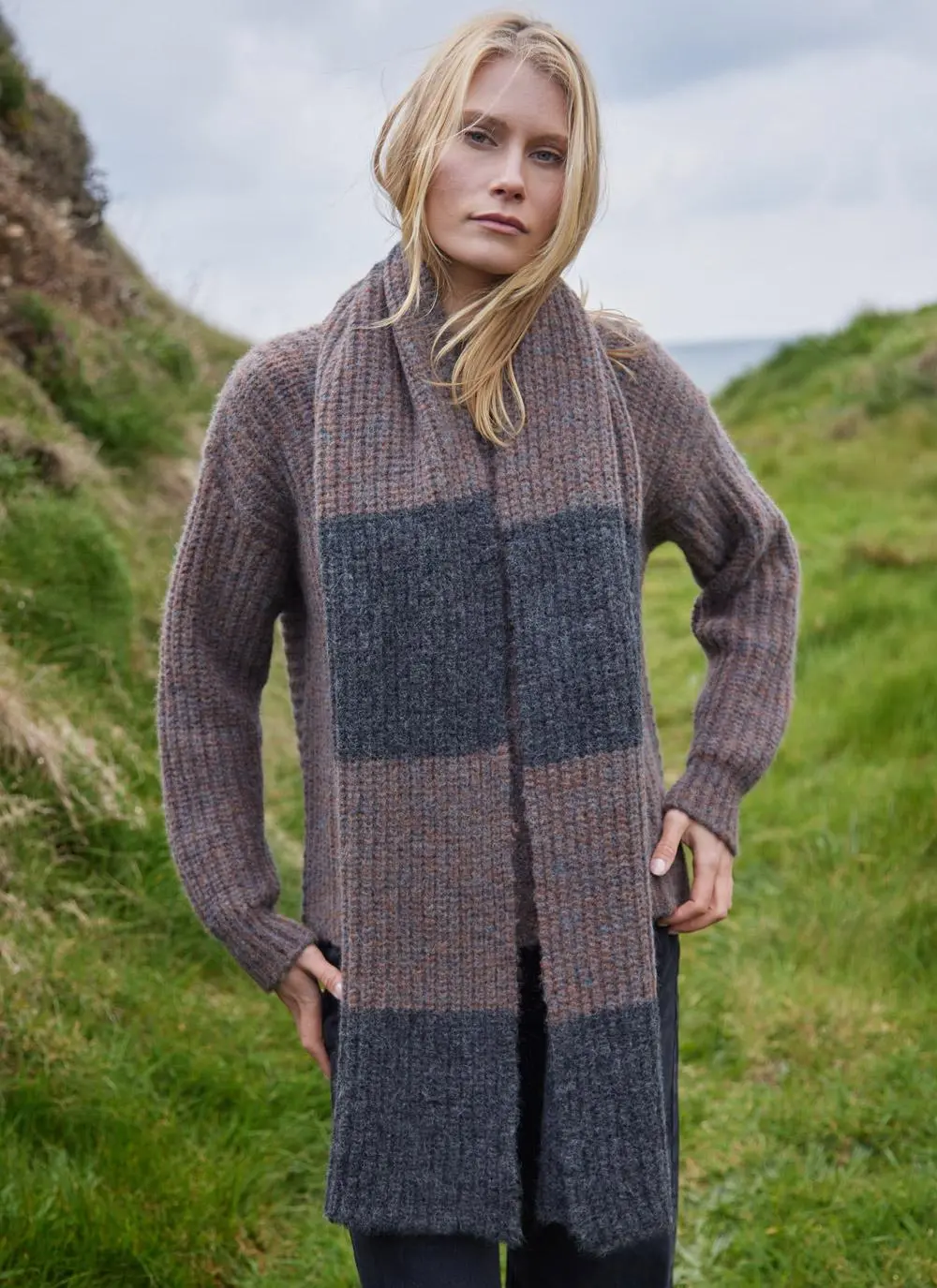 blonde woman standing in grassy coastal area wearing a copper and grey knit scarf with hands near hips