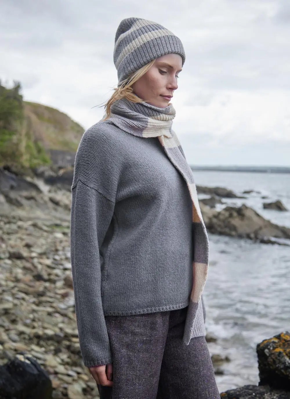 blonde woman wearing grey and white striped hat with a grey sweater and coastal background