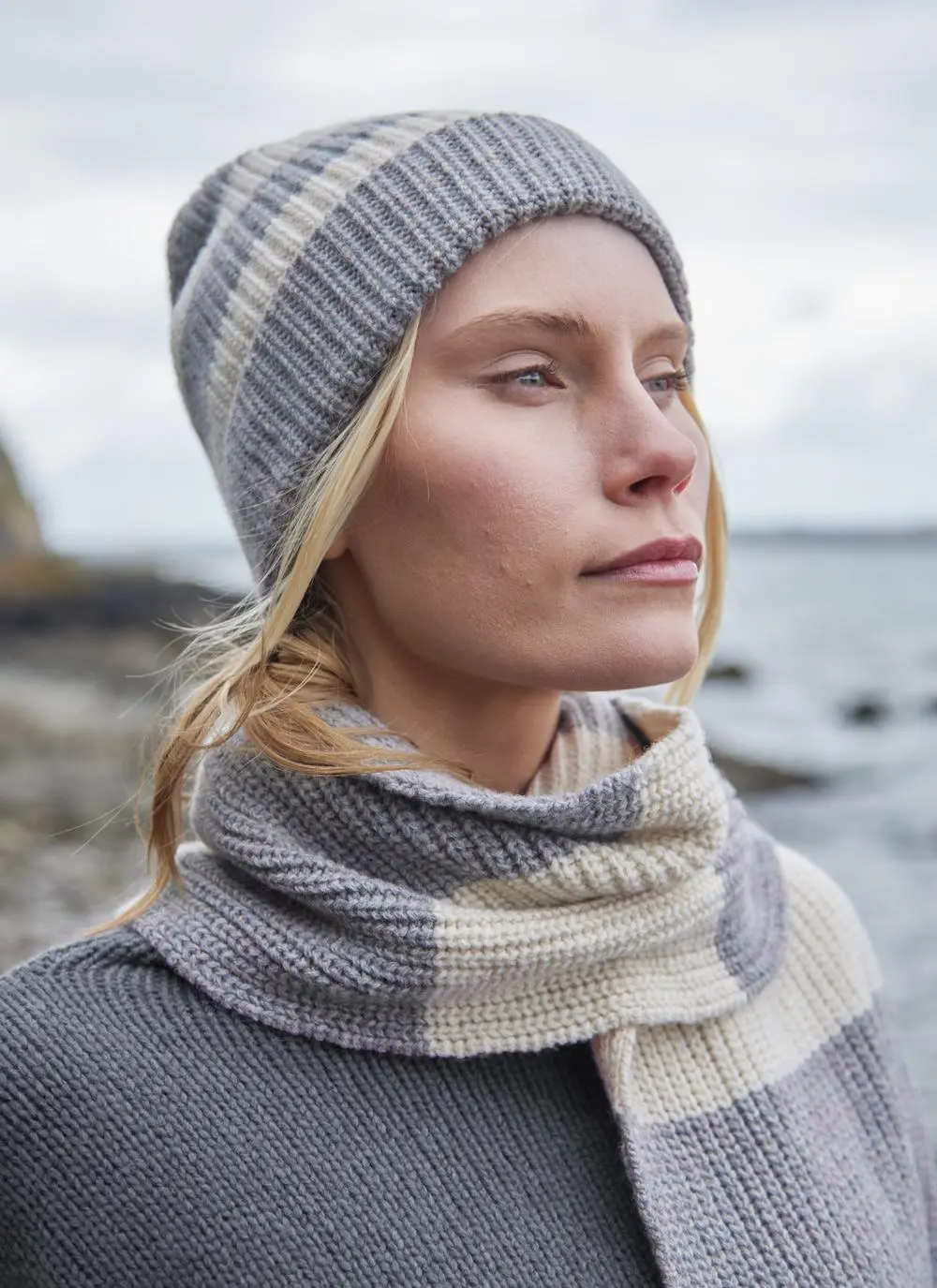 blonde woman wearing grey and white striped hat with a grey sweater and coastal background