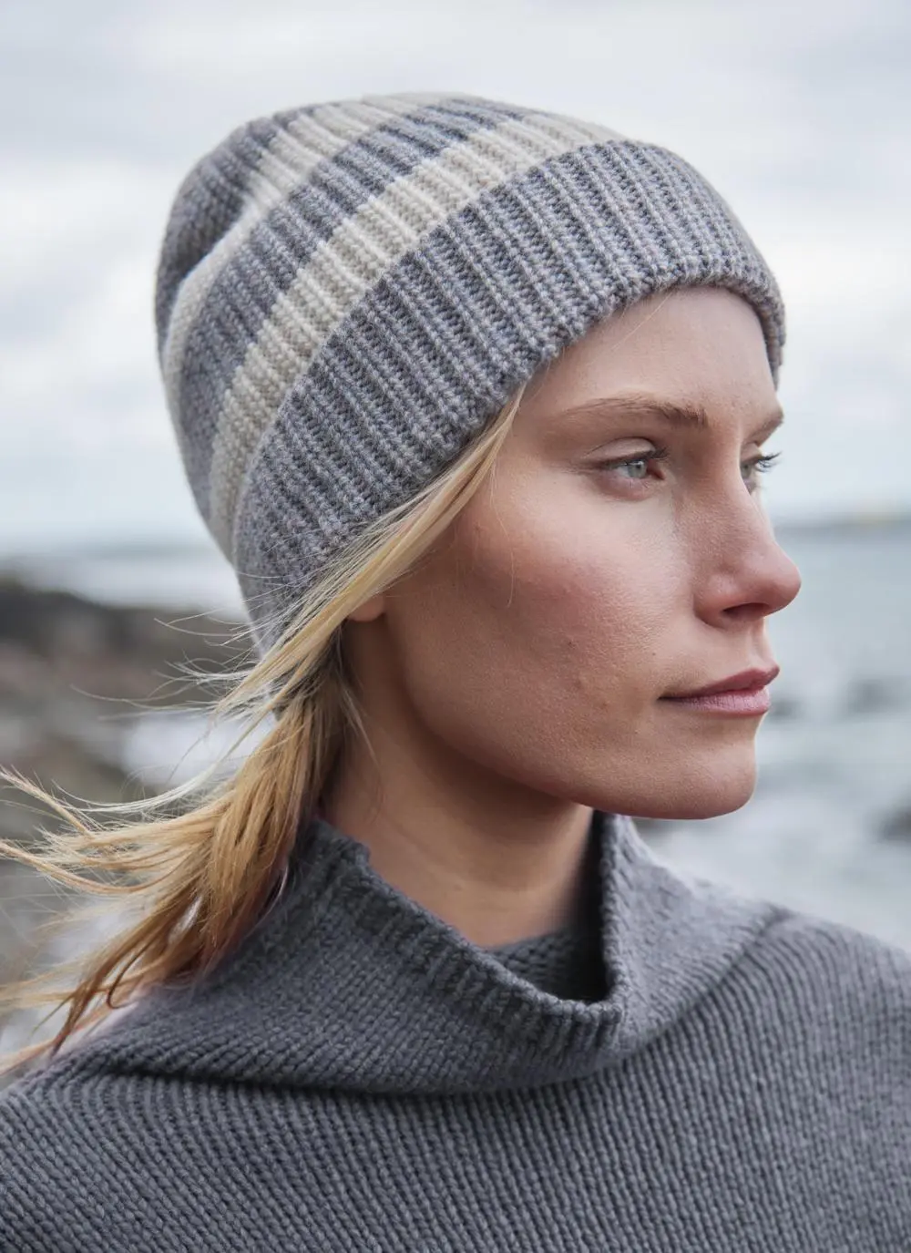 blonde woman wearing grey and white striped hat with a grey sweater and coastal background