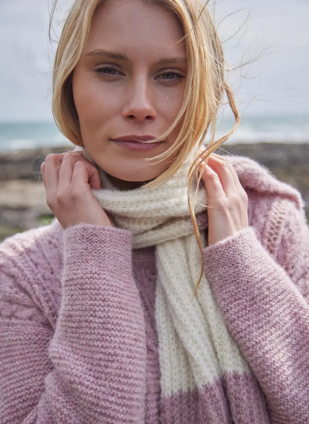 close up of woman blonde woman standing on rocky, mossy beach wearing large knit pink and white scarf