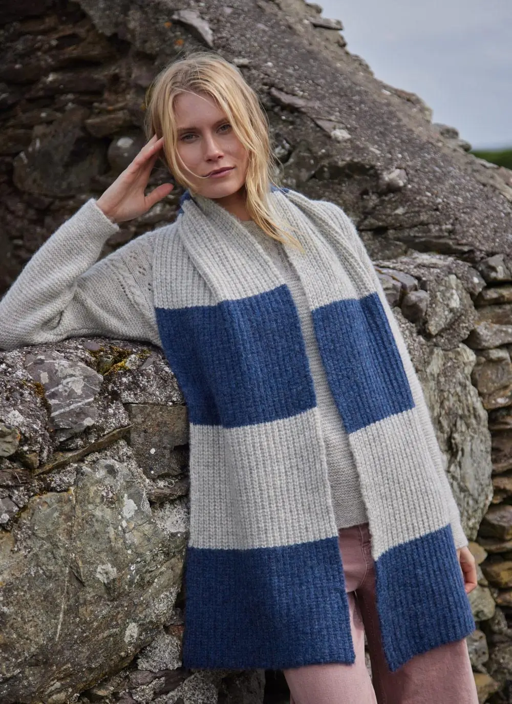 blonde woman leaning against rock wearing a large grey and blue knit scarf 