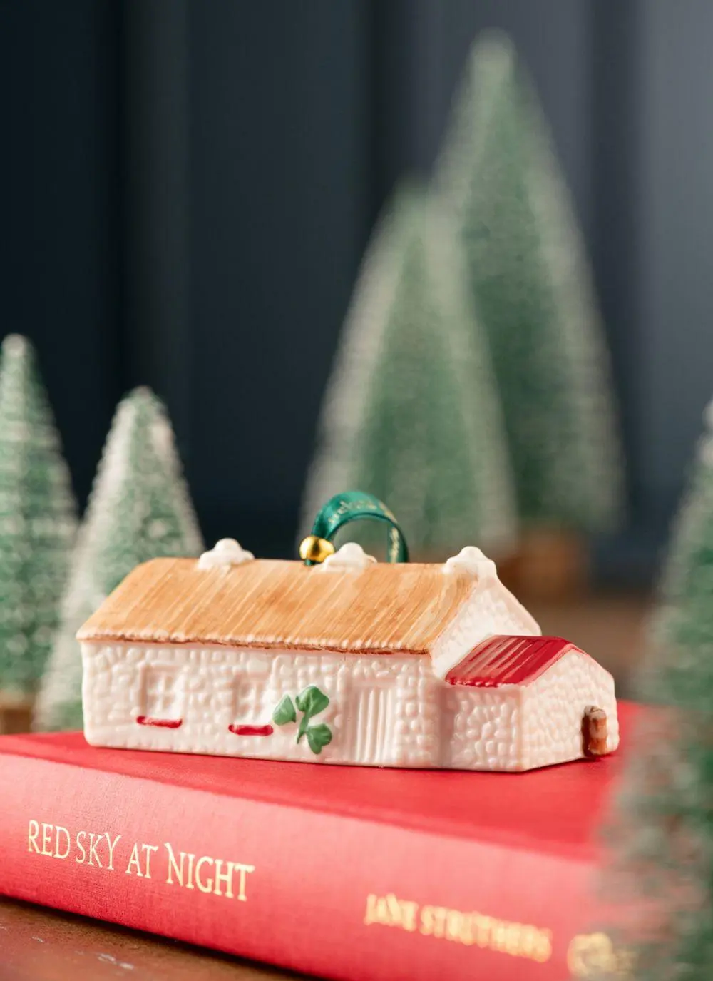 the Belleek Annual Keenaghan Cottage Ornament on a red book with a green shamrock painted on it and red windowsills in front of a background of miniature trees 