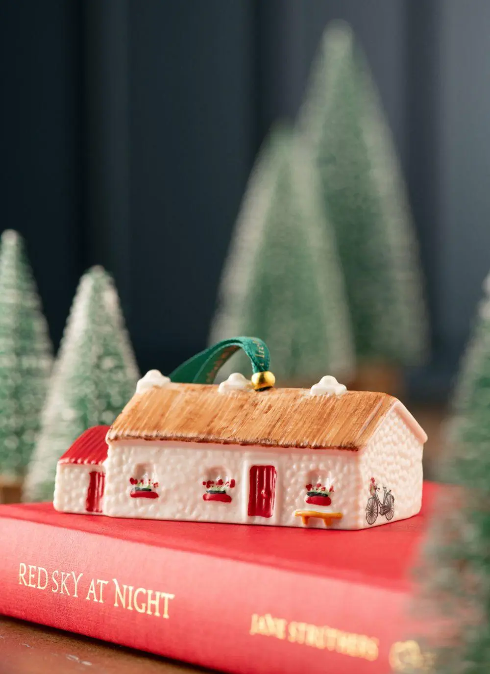 the Belleek Annual Keenaghan Cottage Ornament with a red door, windows a bike and red flowers on the windowsill on a red book in front of a background of miniature trees 