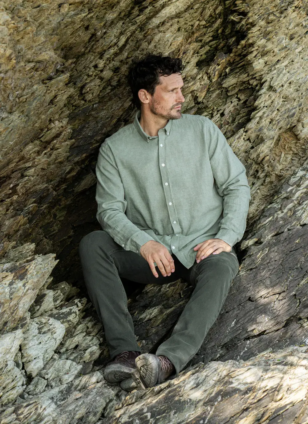 Model seated in a rocky alcove wearing the Finn Cotton Linen Shirt in Green, styled with dark trousers – breathable Irish linen blend.