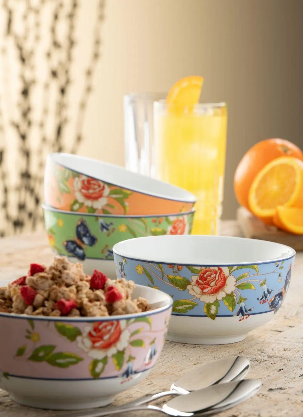 Three floral-patterned bowls, one filled with cereal and berries, on a kitchen counter beside two spoons, with a glass of orange juice and fresh oranges in the background.