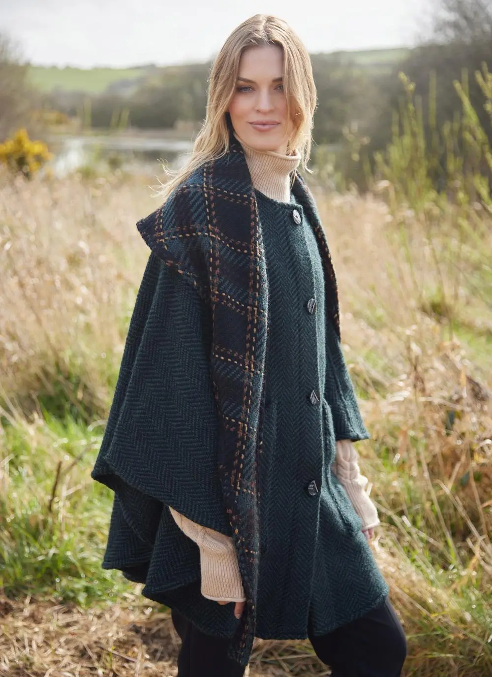 Side angle of blonde woman in grassy Irish countryside wearing dark green herringbone cape.