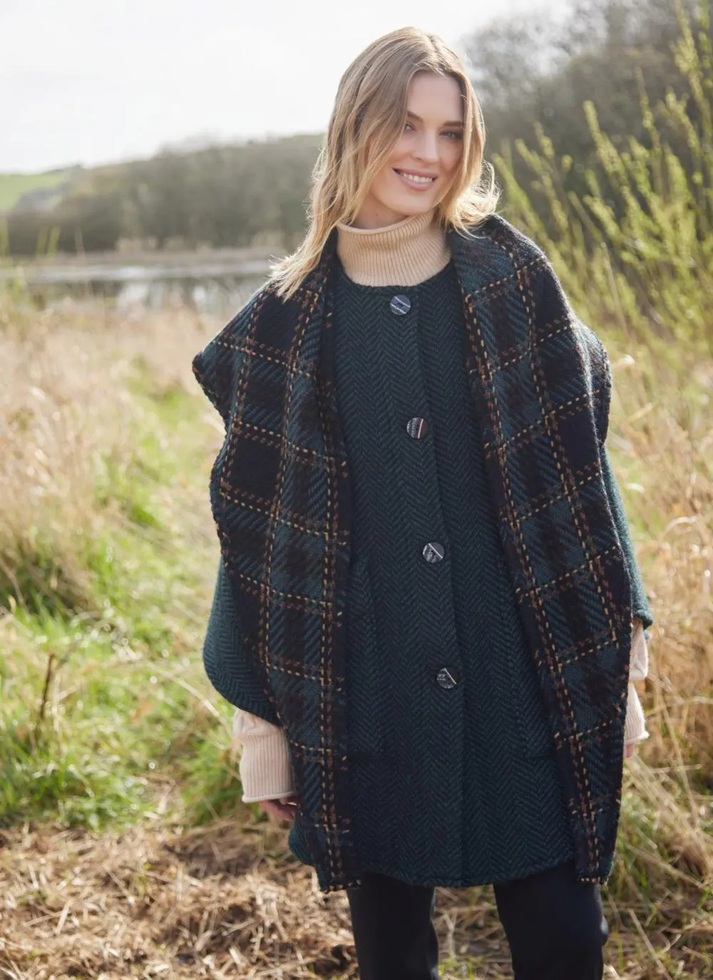 Blonde woman in grassy Irish countryside wearing dark green herringbone cape.