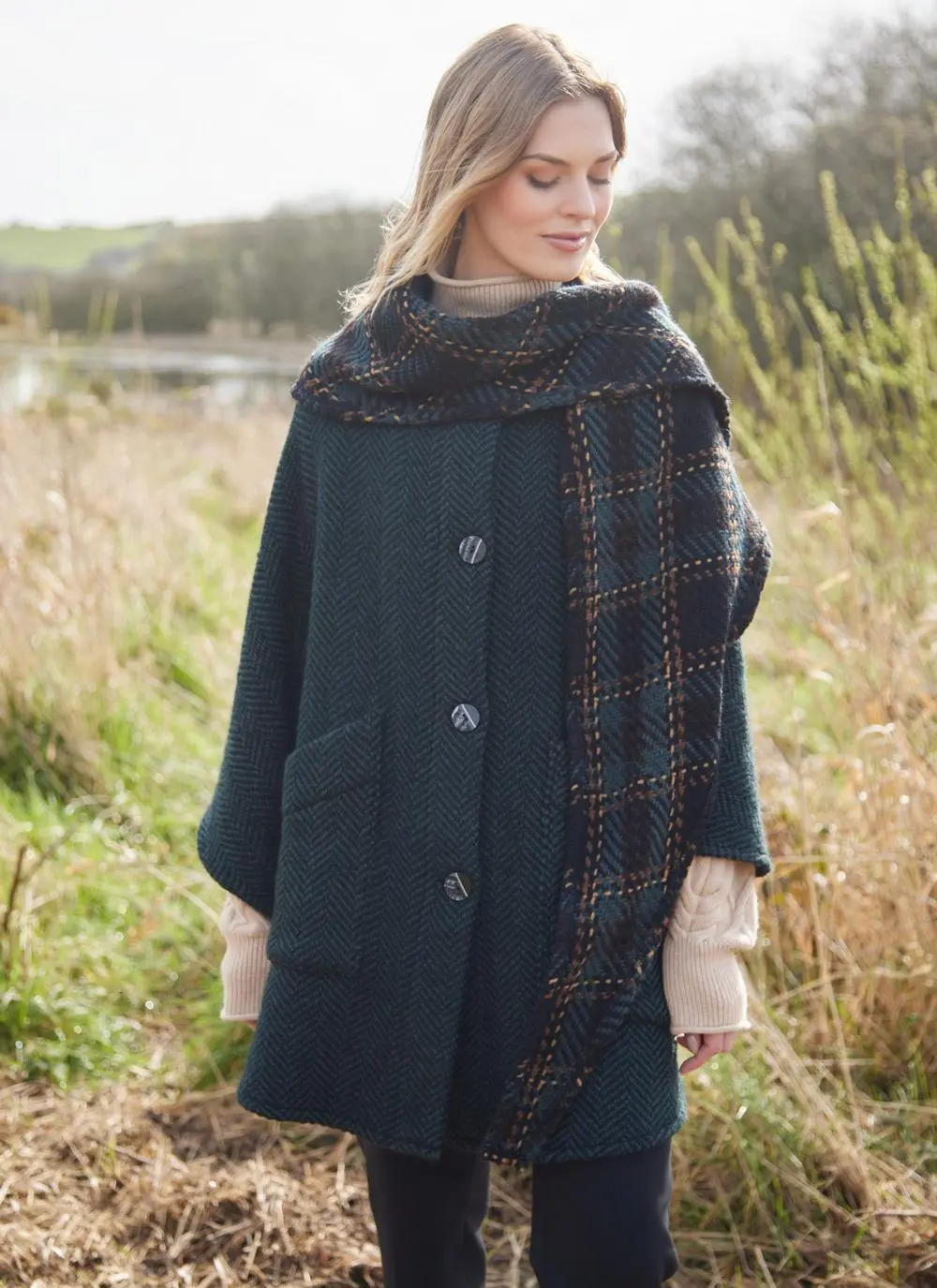Blonde woman in grassy Irish countryside wearing dark green herringbone cape looking to side. 