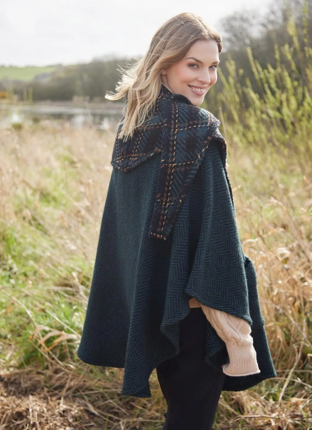 Blonde woman in grassy Irish countryside wearing dark green herringbone cape looking over shoulder. 