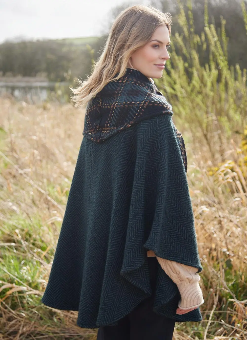 Back angle of blonde woman in grassy Irish countryside wearing dark green herringbone cape.