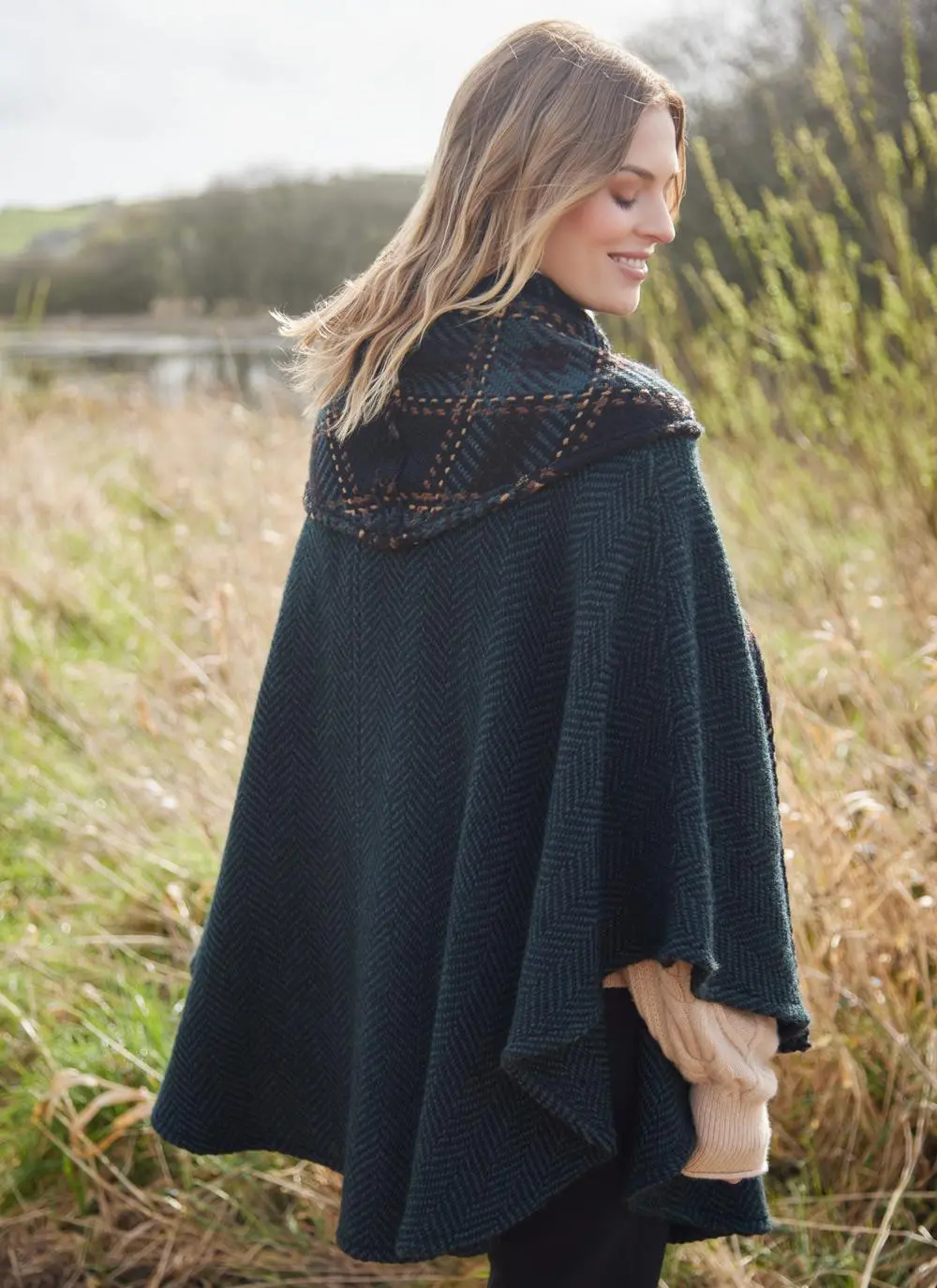 Back angle of blonde woman in grassy Irish countryside wearing dark green herringbone cape.