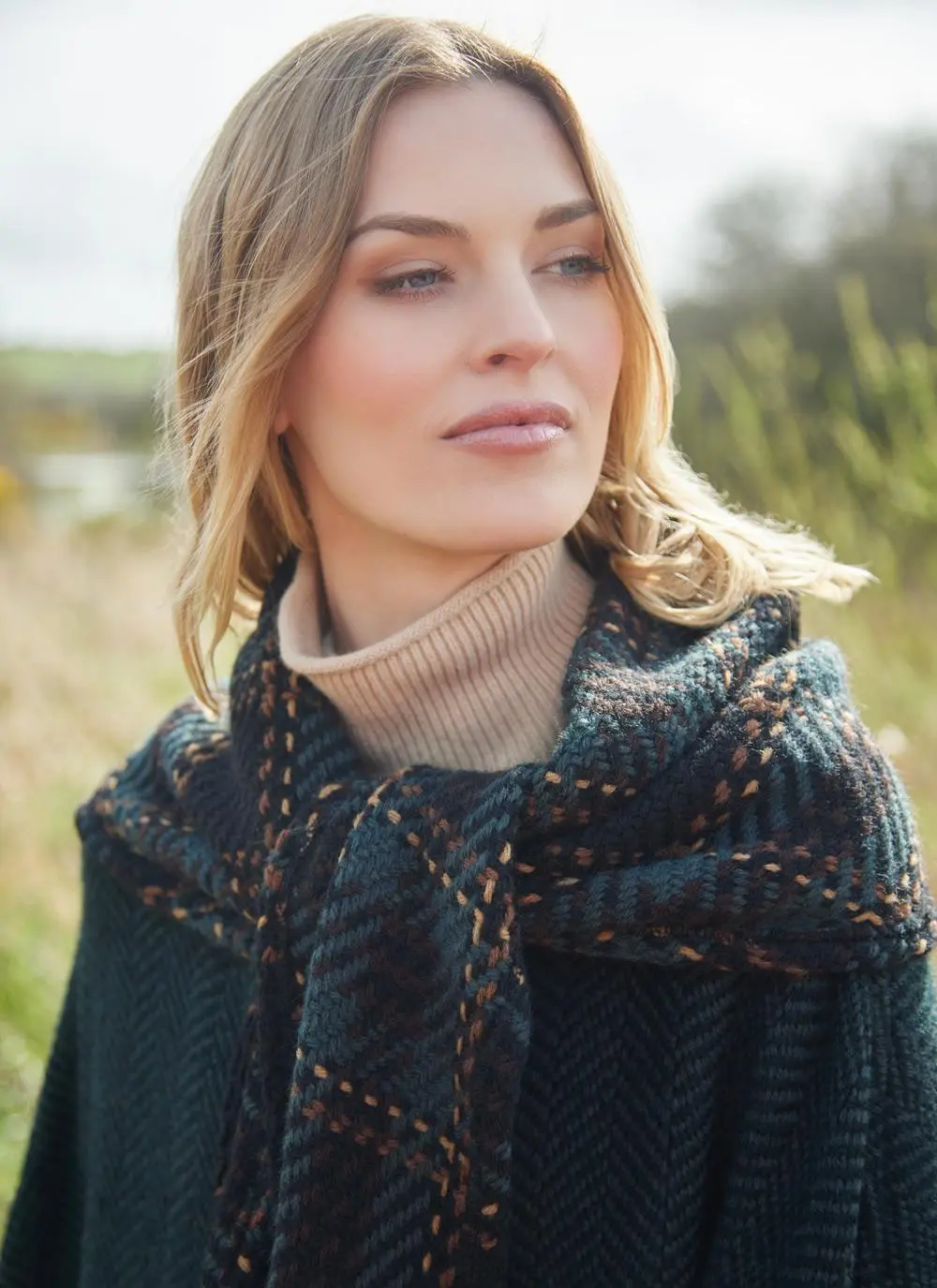 Close up of blonde woman in grassy Irish countryside wearing dark green herringbone cape.