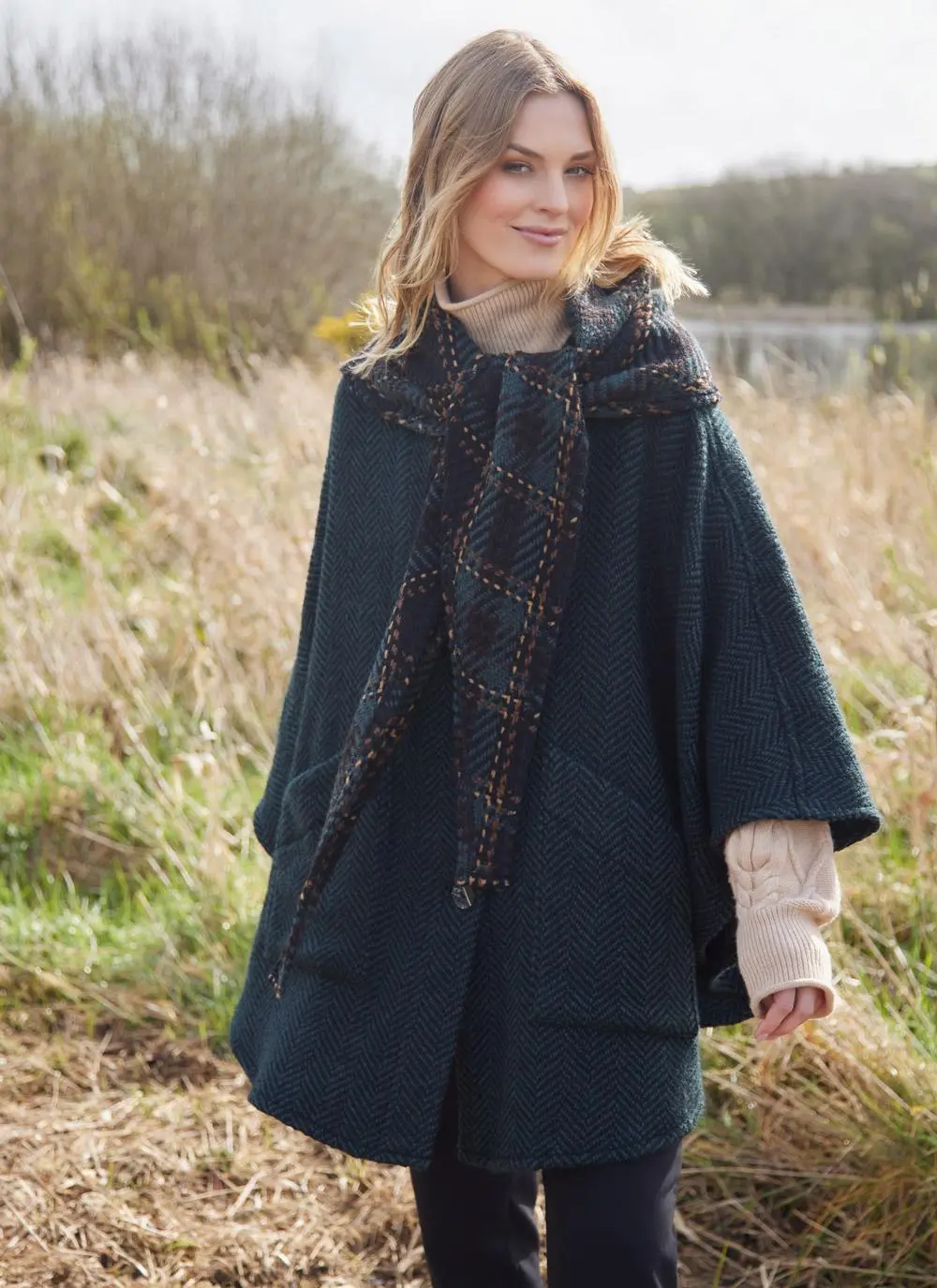 Blonde woman in grassy Irish countryside wearing dark green herringbone cape with hands by side.