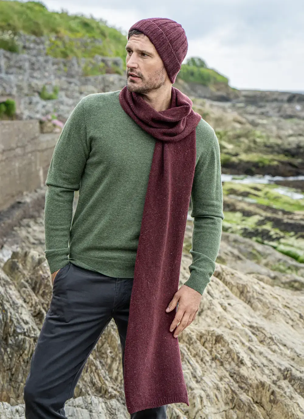 Man wearing a burgundy wool cashmere tweed scarf draped over a green sweater, standing on a rocky coastal path in Ireland.
