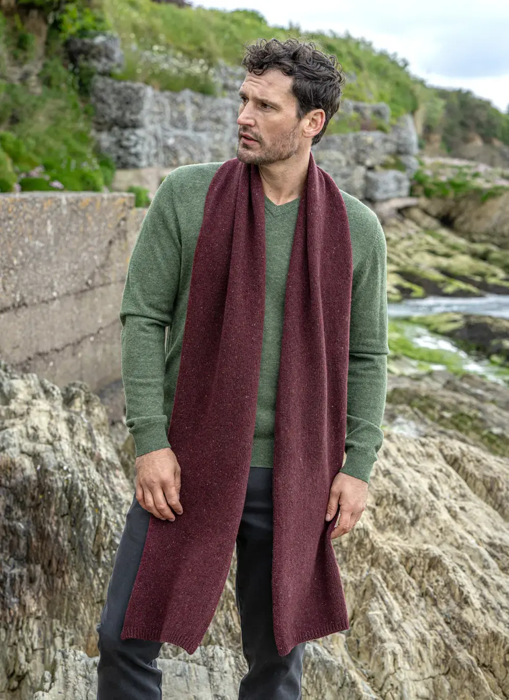 Man wearing a burgundy wool cashmere tweed scarf draped over a green sweater, standing on a rocky coastal path in Ireland.