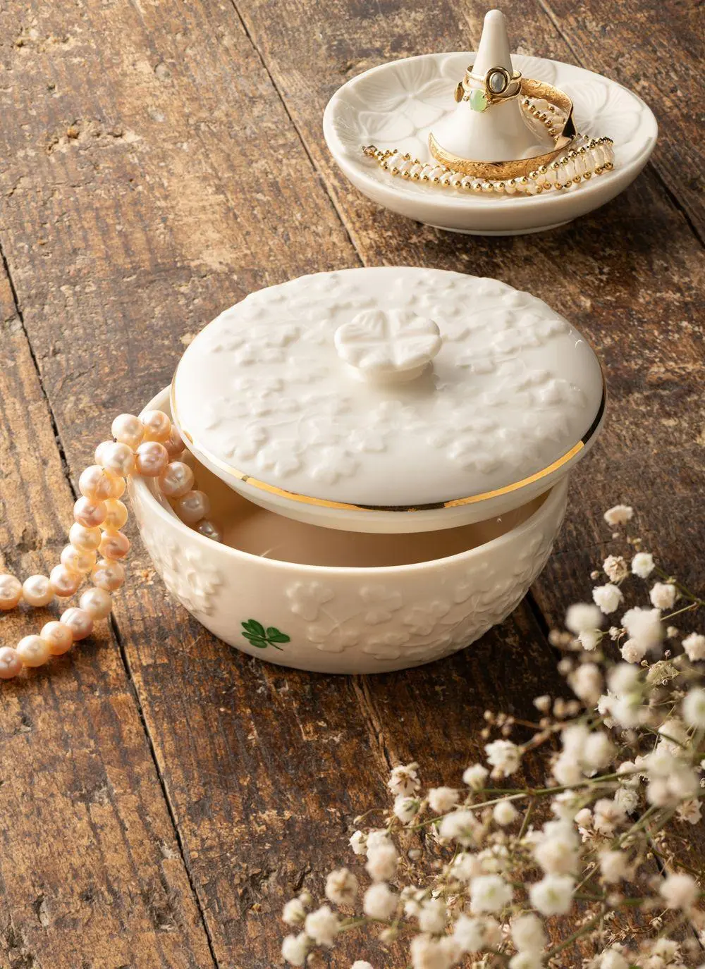 A ceramic jewelry box with a white embossed design sits on a wooden surface, next to a strand of pearls and a plate with rings. Dry flowers add a decorative touch to the rustic setting.