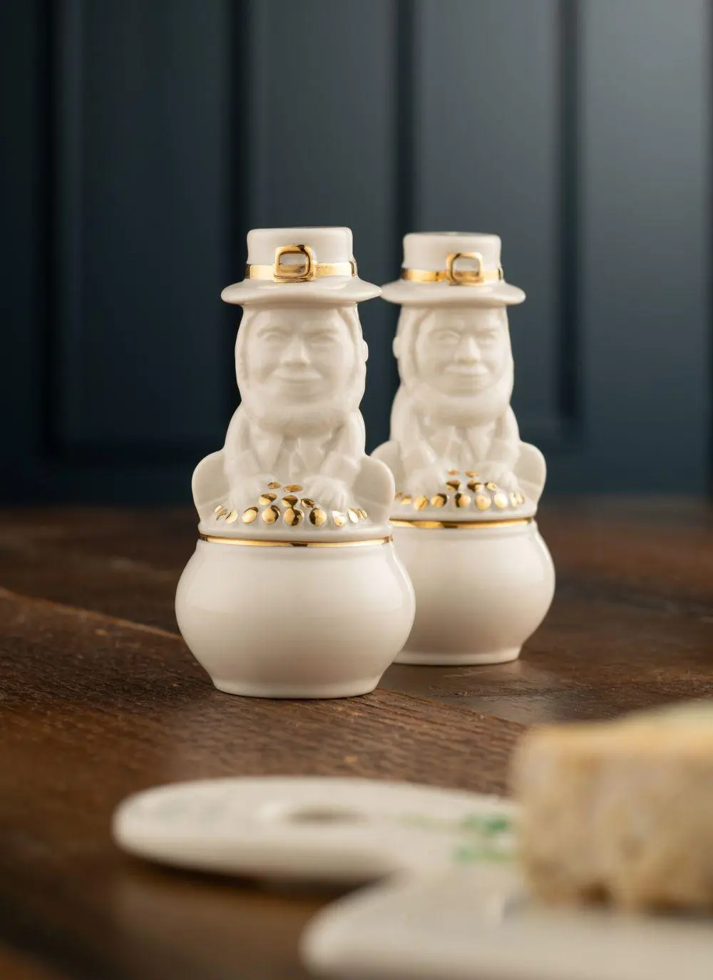 Two white ceramic leprechaun-themed salt and pepper shakers with gold accents, sitting on a wooden table against a dark blue background.