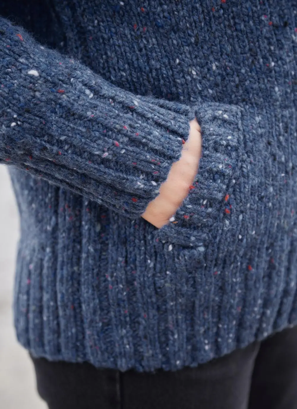 A blue wool cardigan is shown in a close up photograph with a hand in the pocket of the cardigan. 