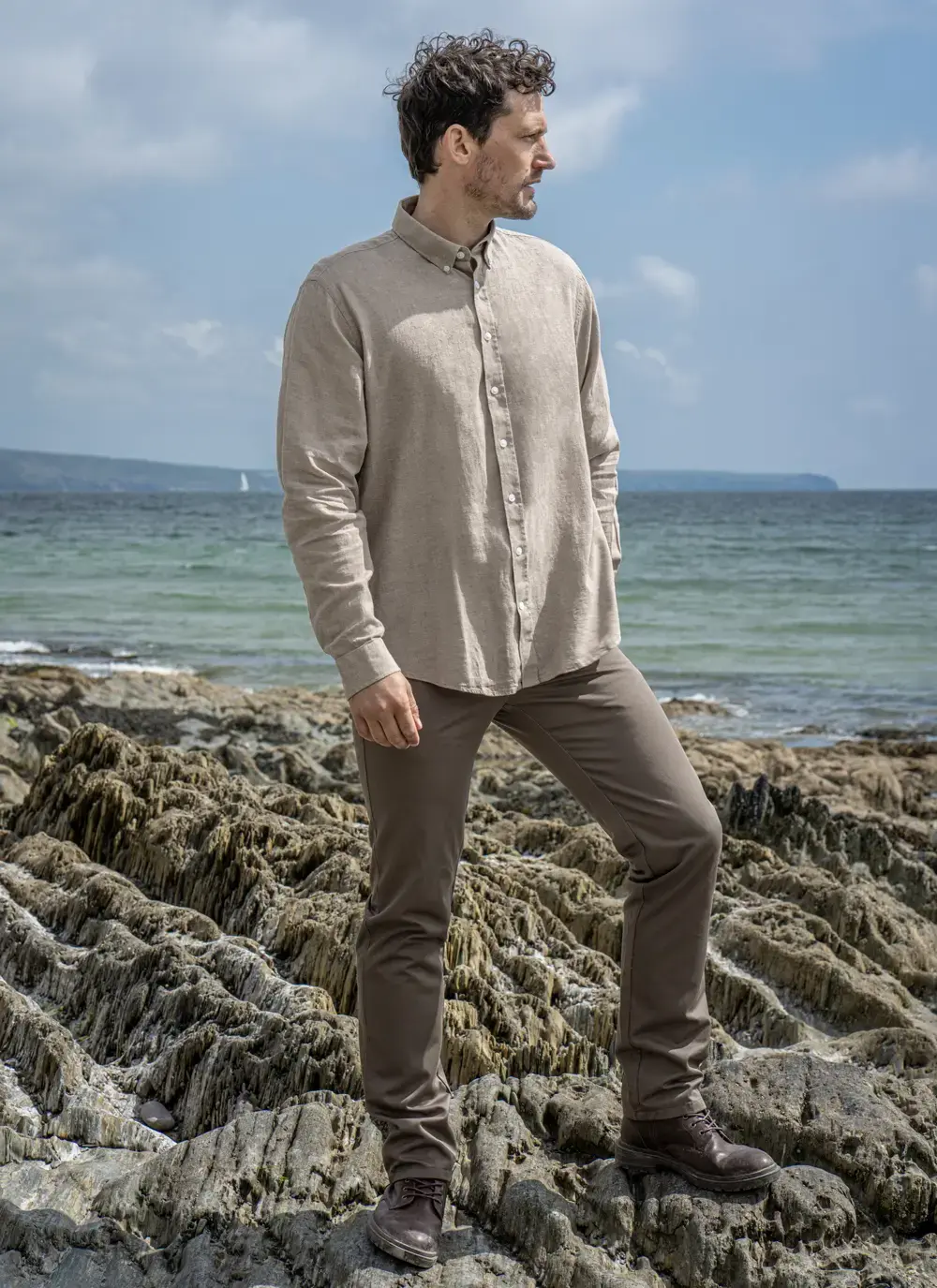 Full body view of man in Finn cotton linen shirt on rocky beach, looking sideways