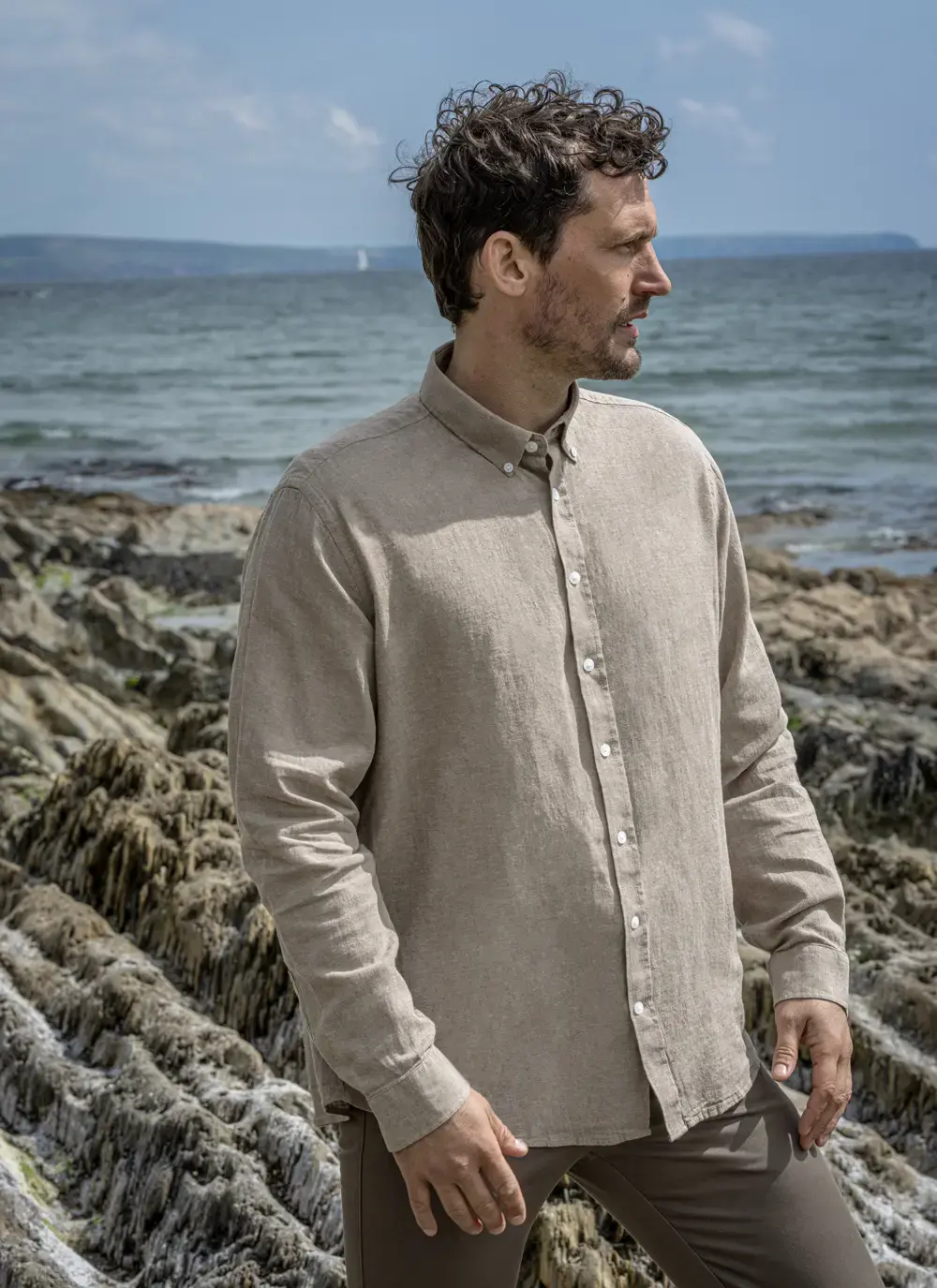 Man wearing Finn cotton linen shirt standing on rocky seashore, looking up, front view