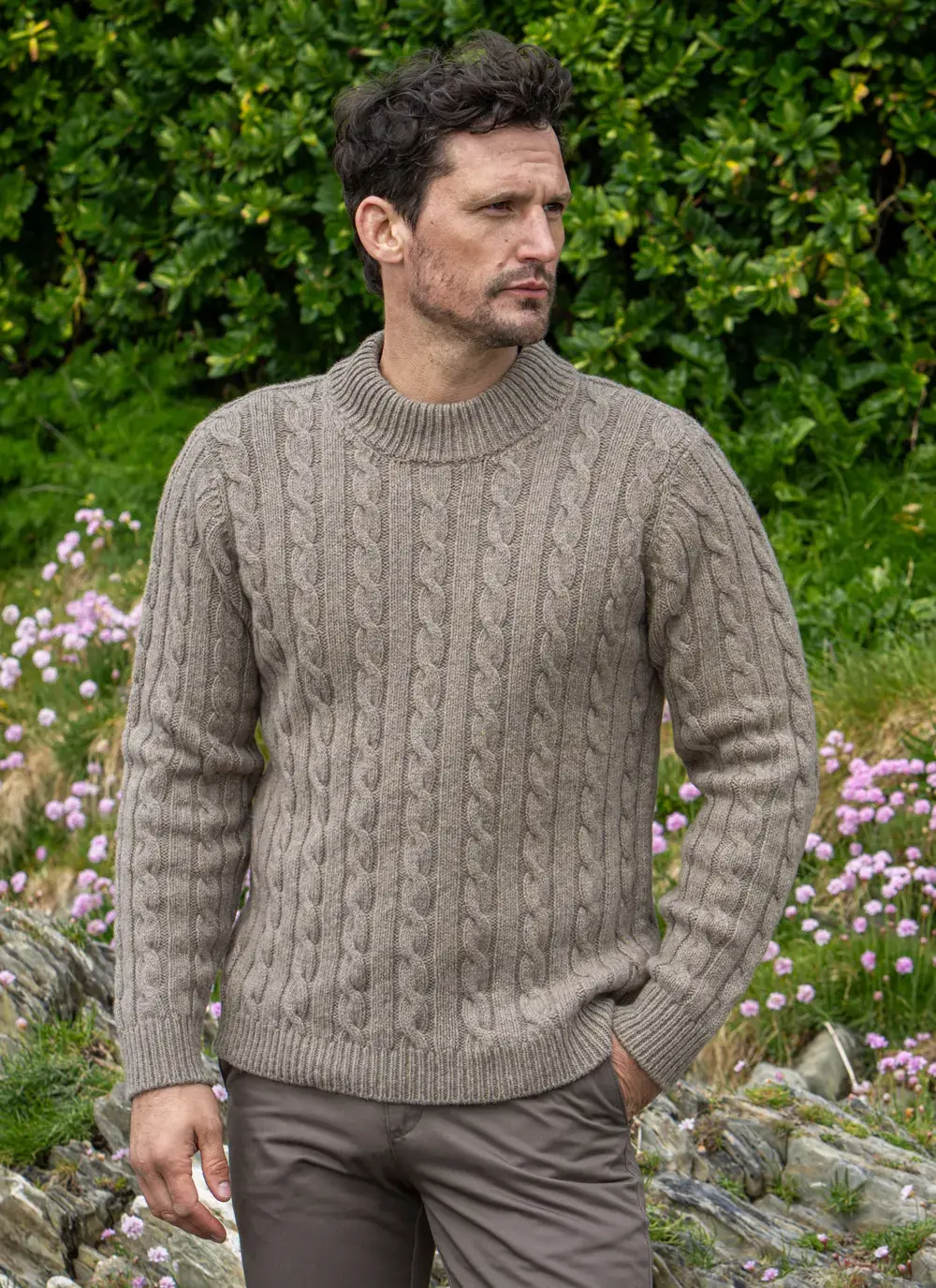 Man wearing Fisherman Cashmere Wool Cable Mock Neck Sweater in Clay, standing outdoors with green foliage and pink wildflowers in the background.