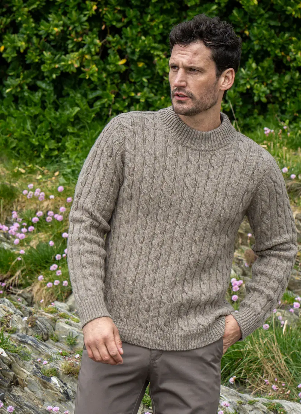 Model wearing Cashmere Wool Fisherman Sweater in Clay, looking away with natural greenery and wildflowers behind.