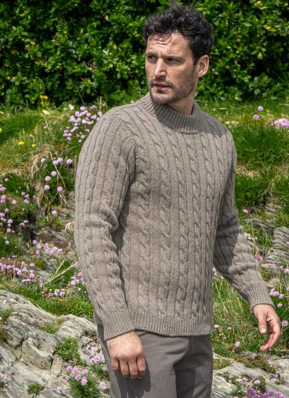 Side view of man in Clay-colored Fisherman Cable Sweater, standing on rocky terrain with pink flowers and greenery.