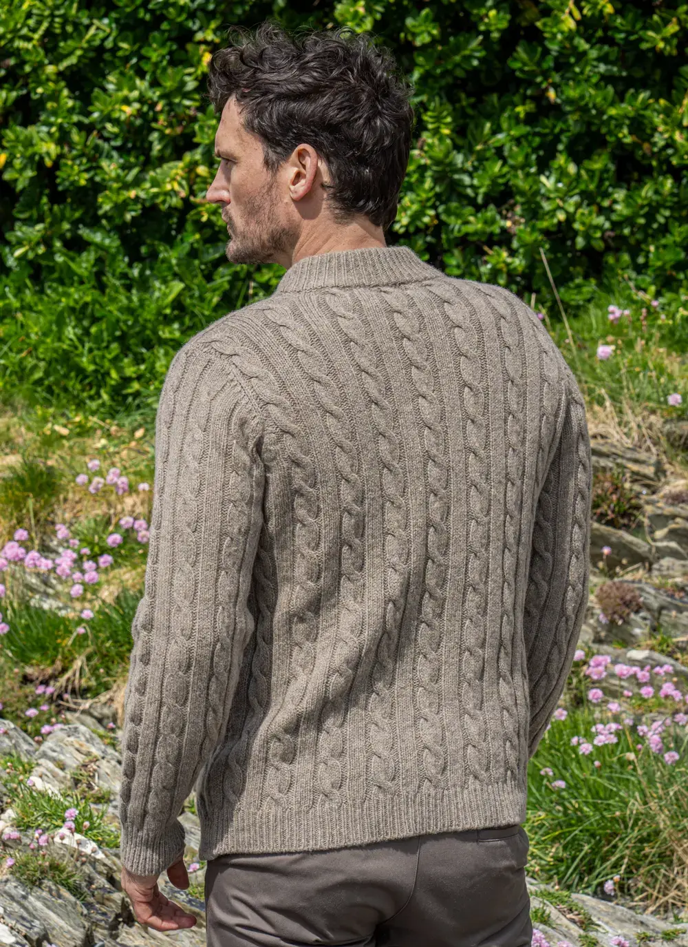 Back view of Fisherman Cable Knit Sweater in Clay, showing full cable pattern and ribbed cuffs, modeled in a natural setting.