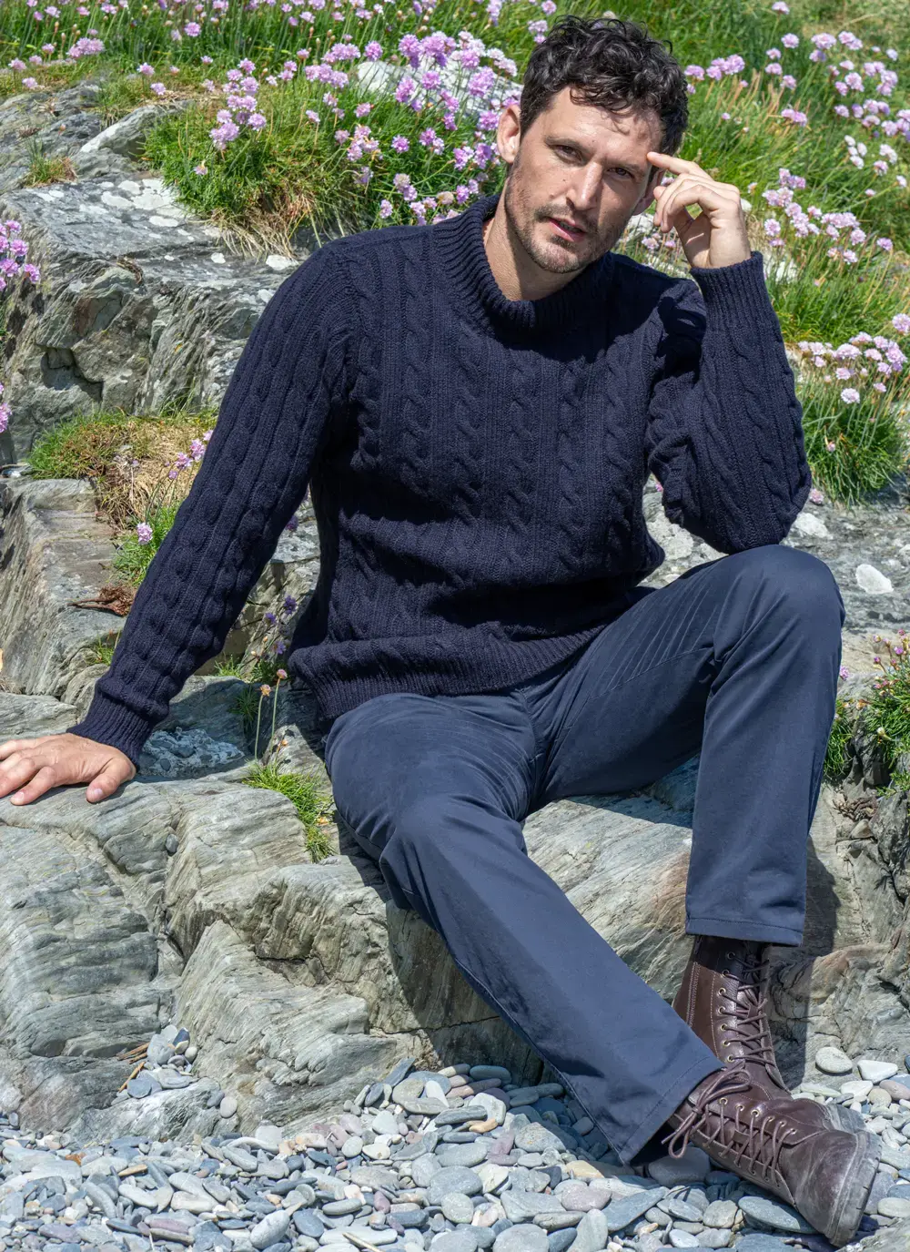 Man sitting on coastal rocks wearing Fisherman Cashmere Wool Cable Mock Neck Sweater in Navy, paired with navy trousers and brown lace-up boots.