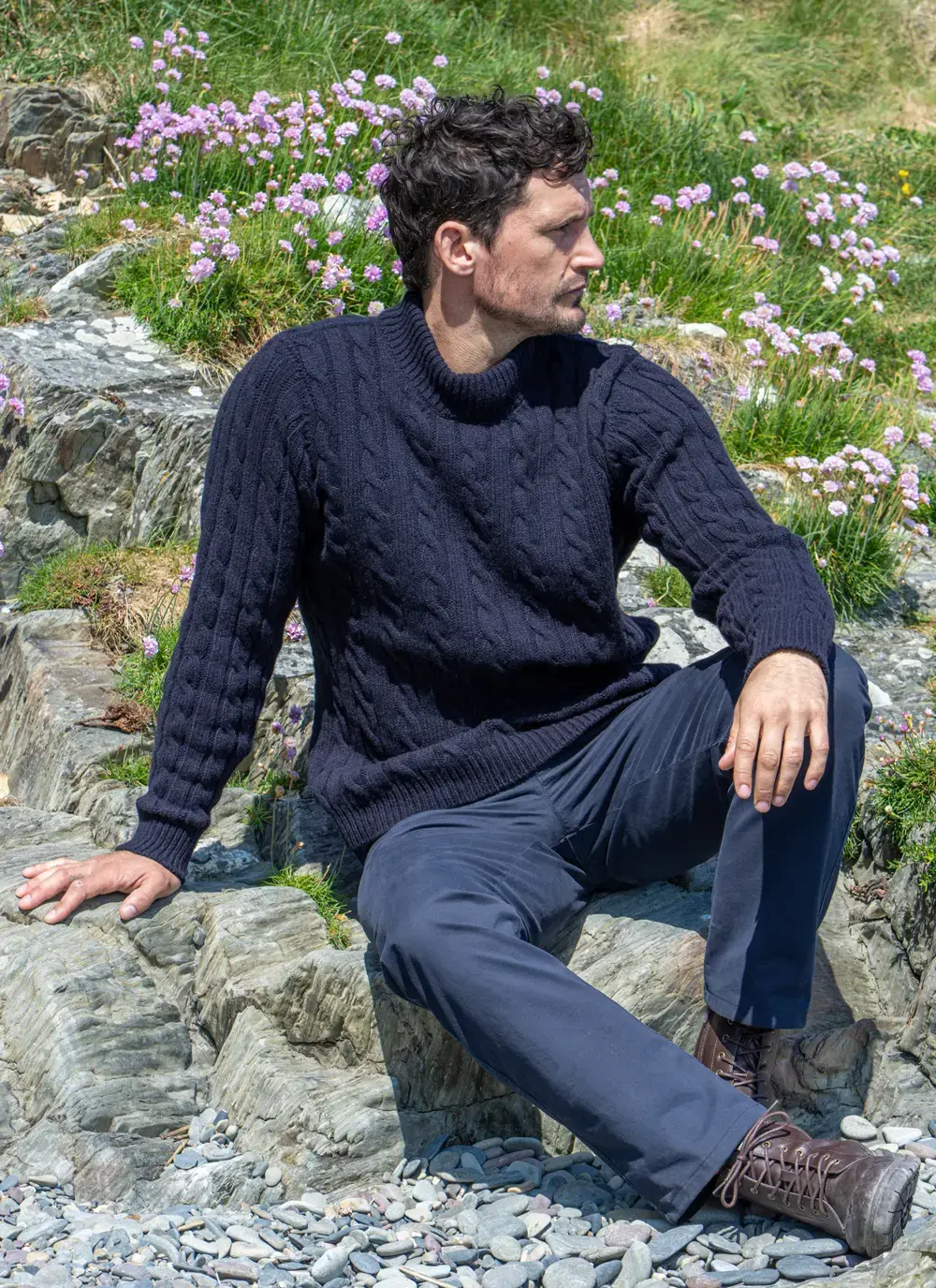 Model seated outdoors in navy Fisherman Cable Knit Sweater, surrounded by wildflowers and rocky terrain.