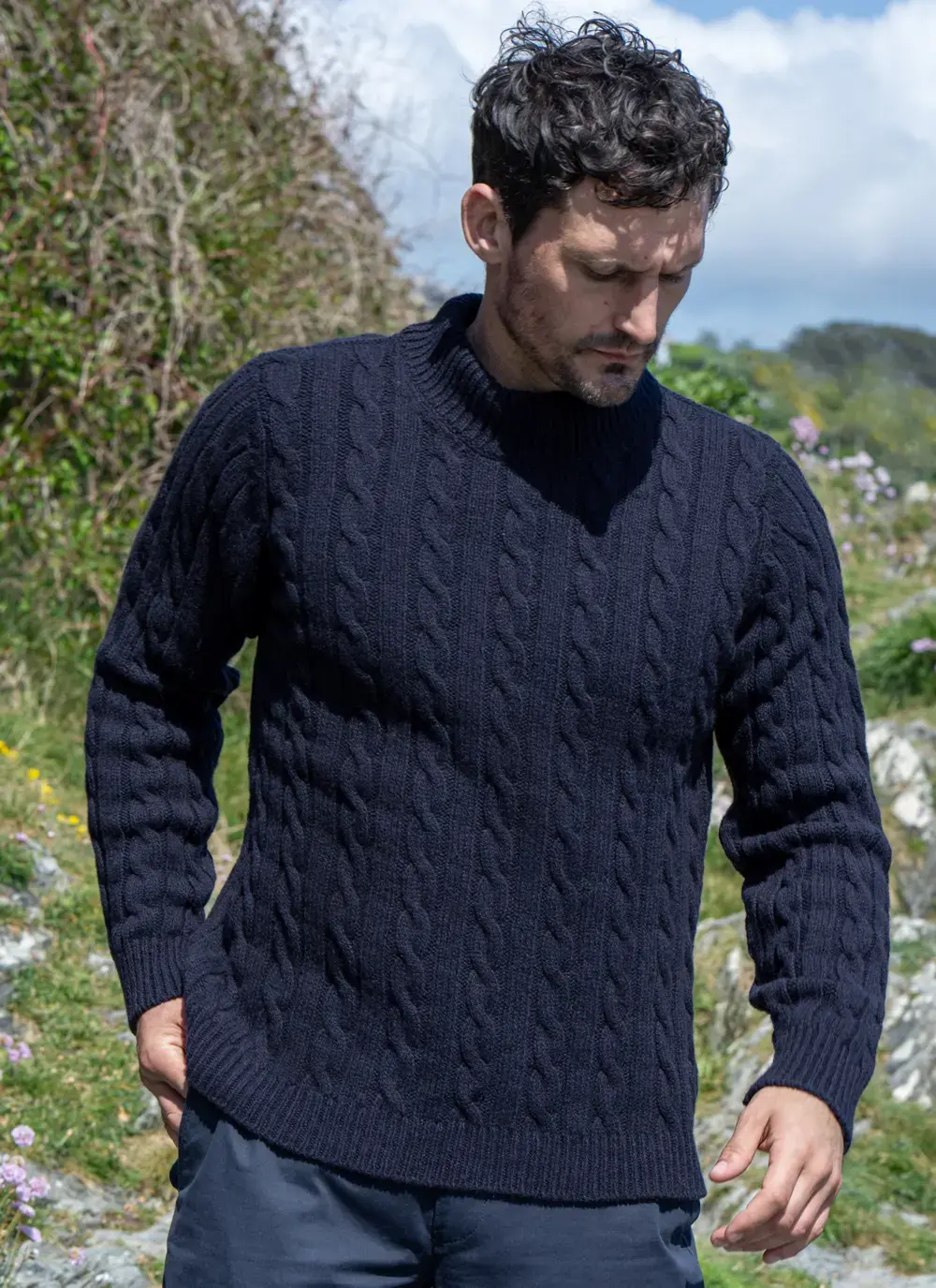 Man looking down while wearing navy cable knit mock neck sweater, with greenery and wildflowers in the background.