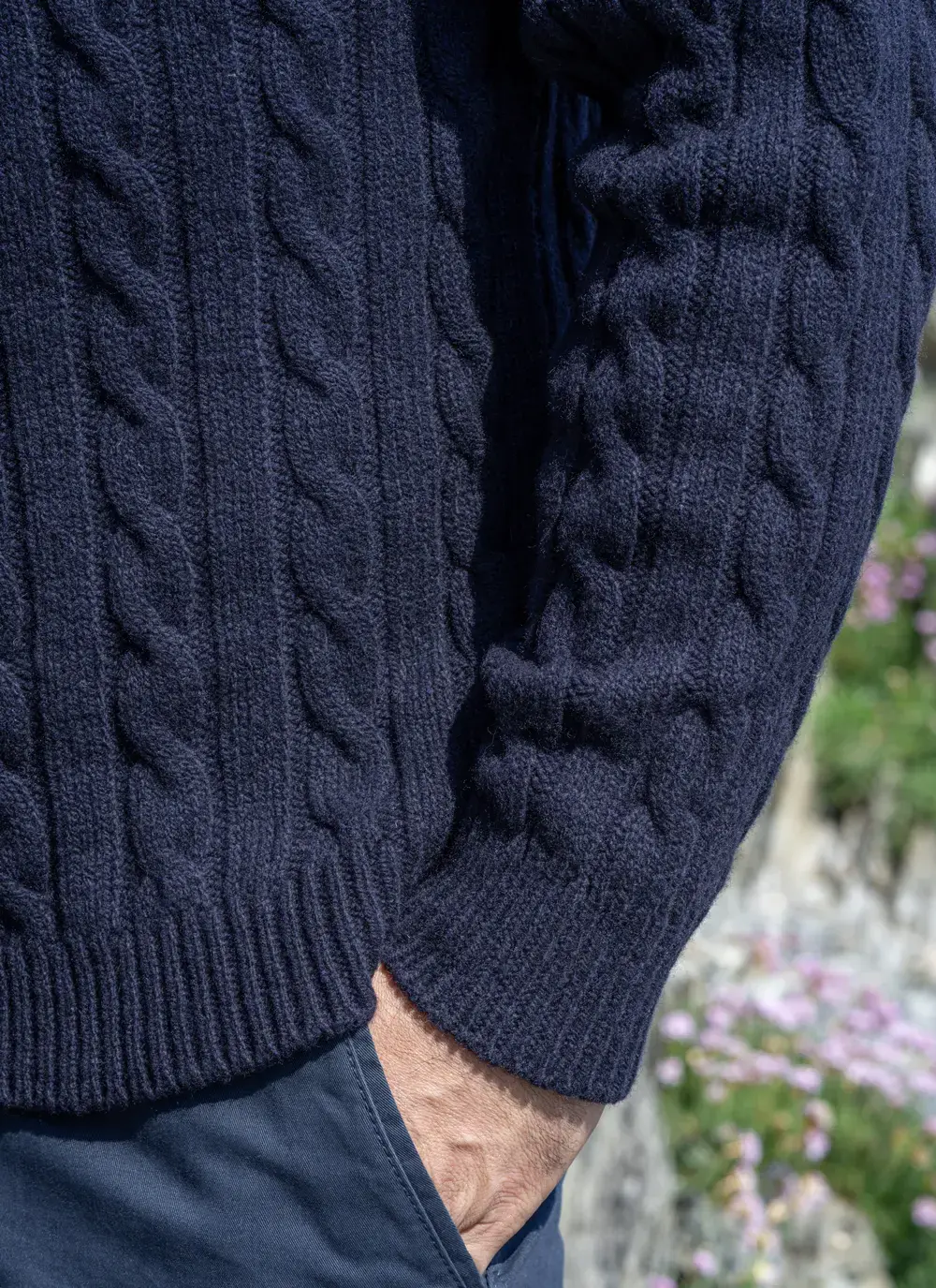 Detailed view of sleeve and hem on navy cable knit sweater, with hand in pocket and natural background.
