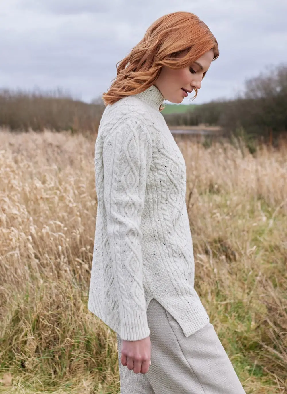 Side shot of red haired woman in field wearing ecru colored Aran turtle neck sweater