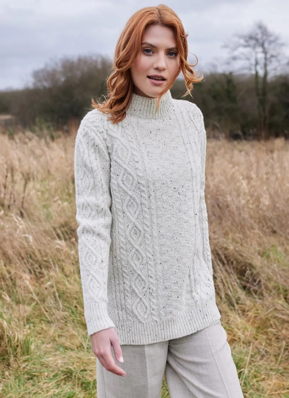 Red haired woman in field wearing ecru colored Aran turtle neck sweater walking