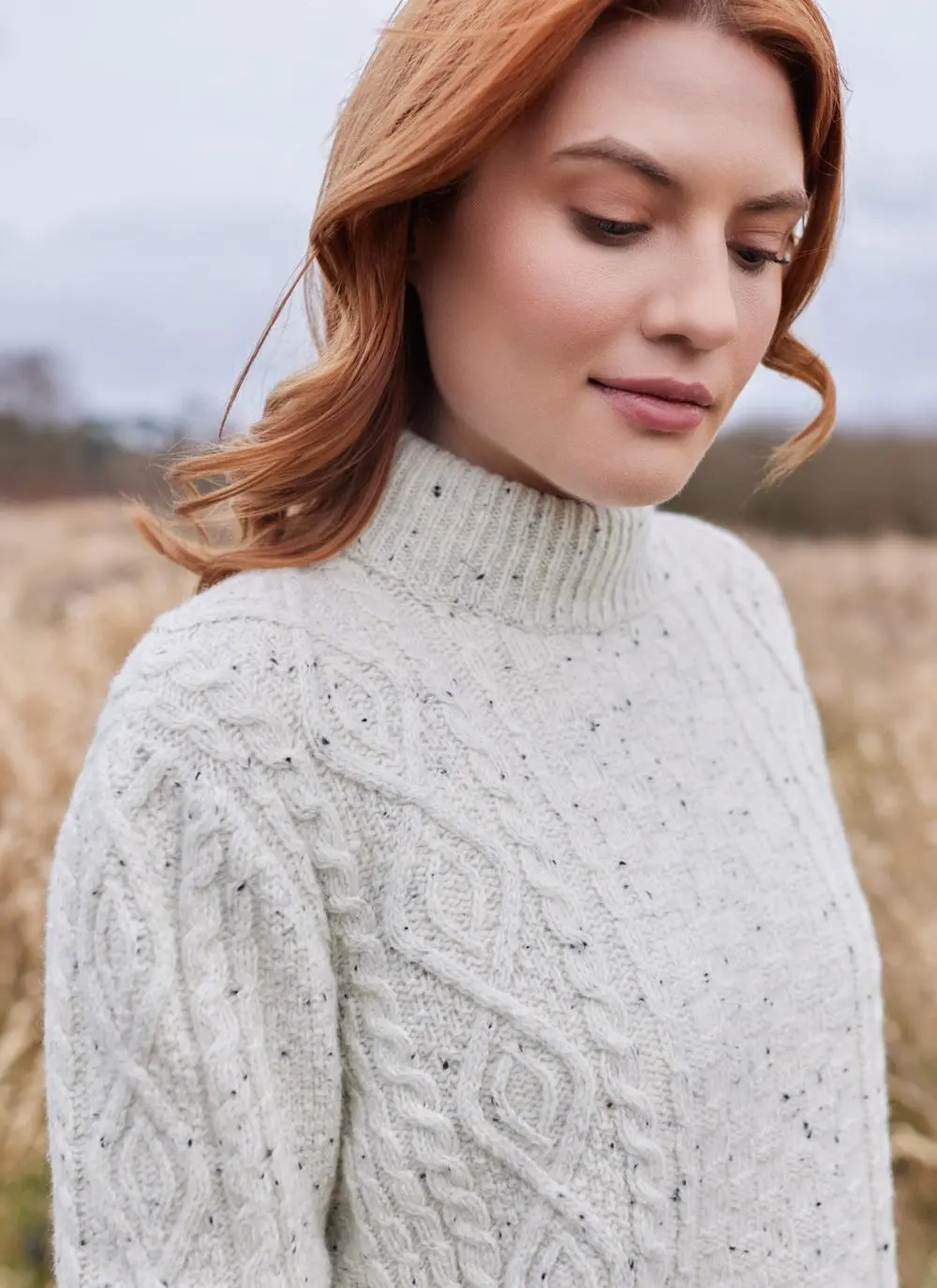 Close up shot of red haired woman in field wearing ecru colored Aran turtle neck sweater
