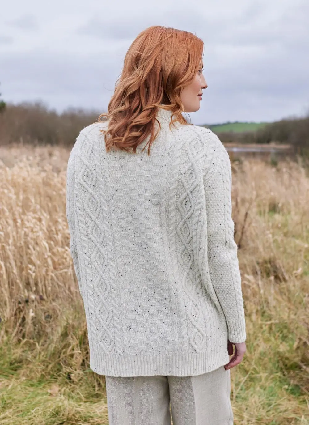 Back shot of red haired woman in field wearing ecru colored Aran turtle neck sweater